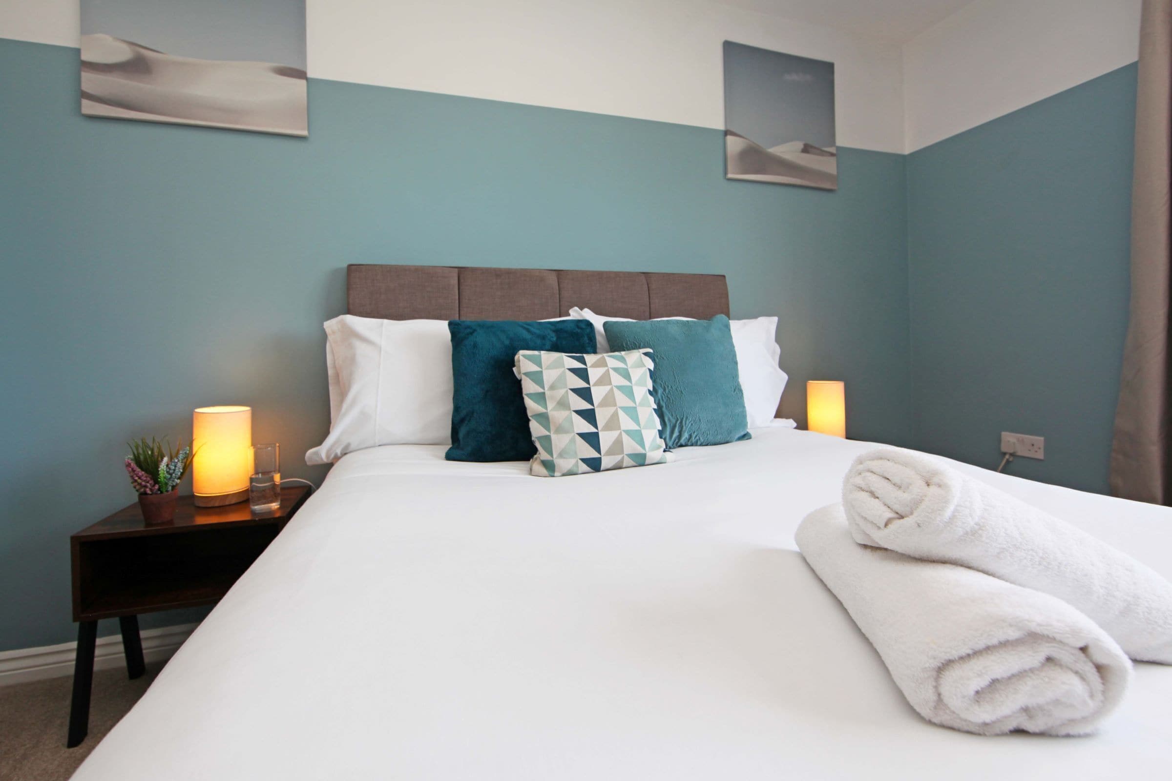A neatly made bed with white and teal pillows, a bedside lamp, and a rolled white towel on the bed.