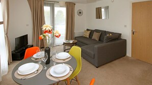 A living room with a grey sofa, a dining table set with plates and glasses, a vase with flowers, and a TV in the corner. - Autumn Heights Apartments (Telford)