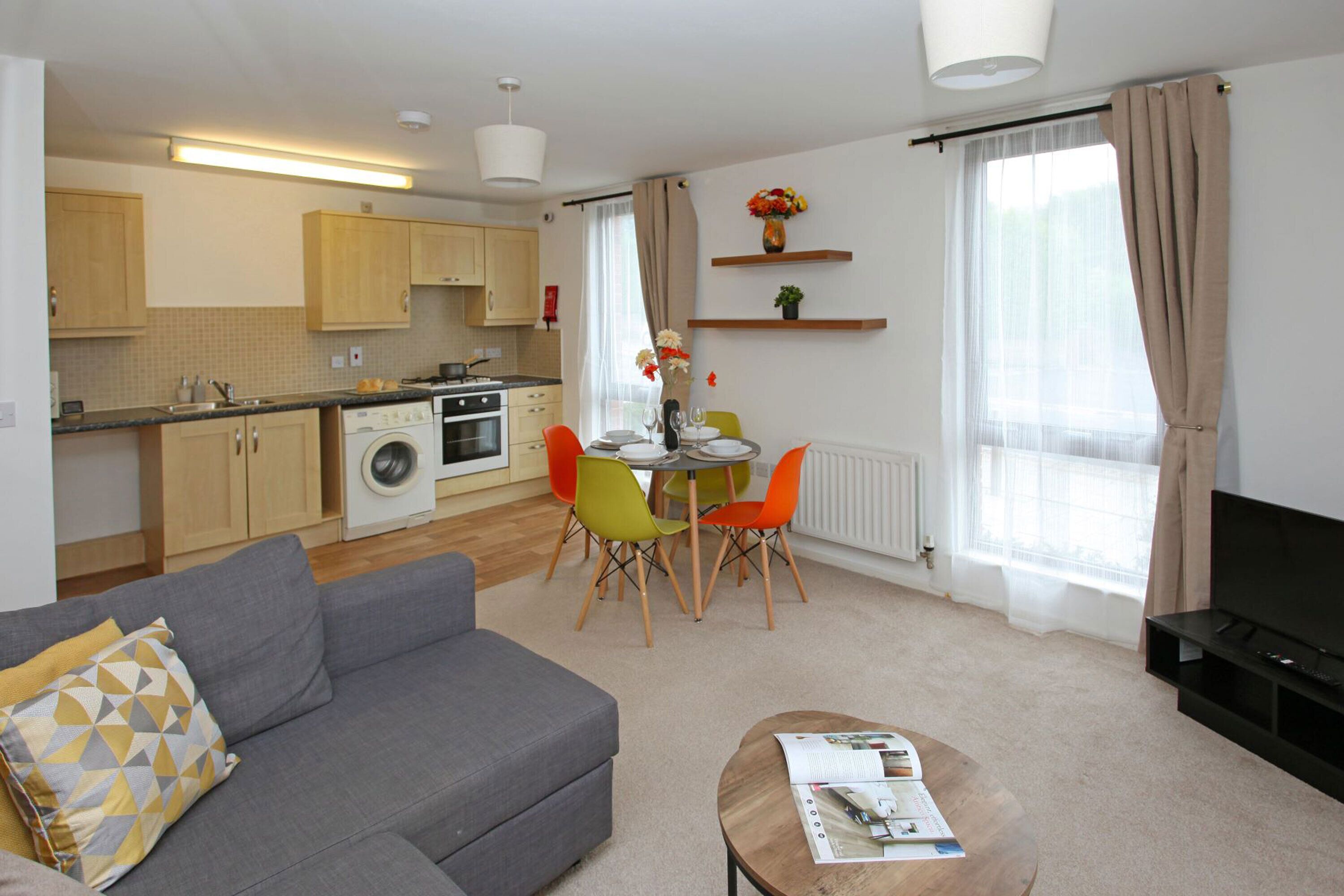 A modern living room with a grey sofa, a coffee table with magazines, a dining area with a table and chairs, a kitchen with wooden cabinets and appliances, and a TV on a stand.