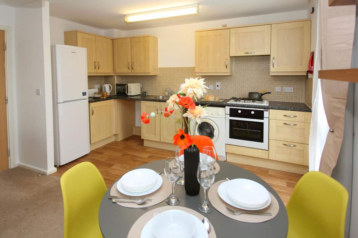 A modern kitchen with wooden cabinets, a refrigerator, microwave, oven, and a dining table set for four with chairs.