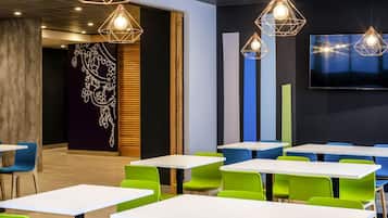 A modern dining area with white tables and green chairs, geometric pendant lighting, and a large flat-screen TV on the wall.