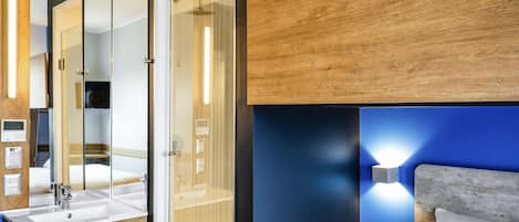 Habitación de hotel moderna con cabina de ducha de vidrio, tocador de madera con lavamanos y cama con cabecero.