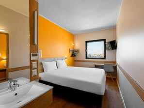 A hotel room with a bed, a desk, a chair, a large window, and a bathroom with a sink and mirror. - Ibis Budget Santiago Providencia (Santiago)