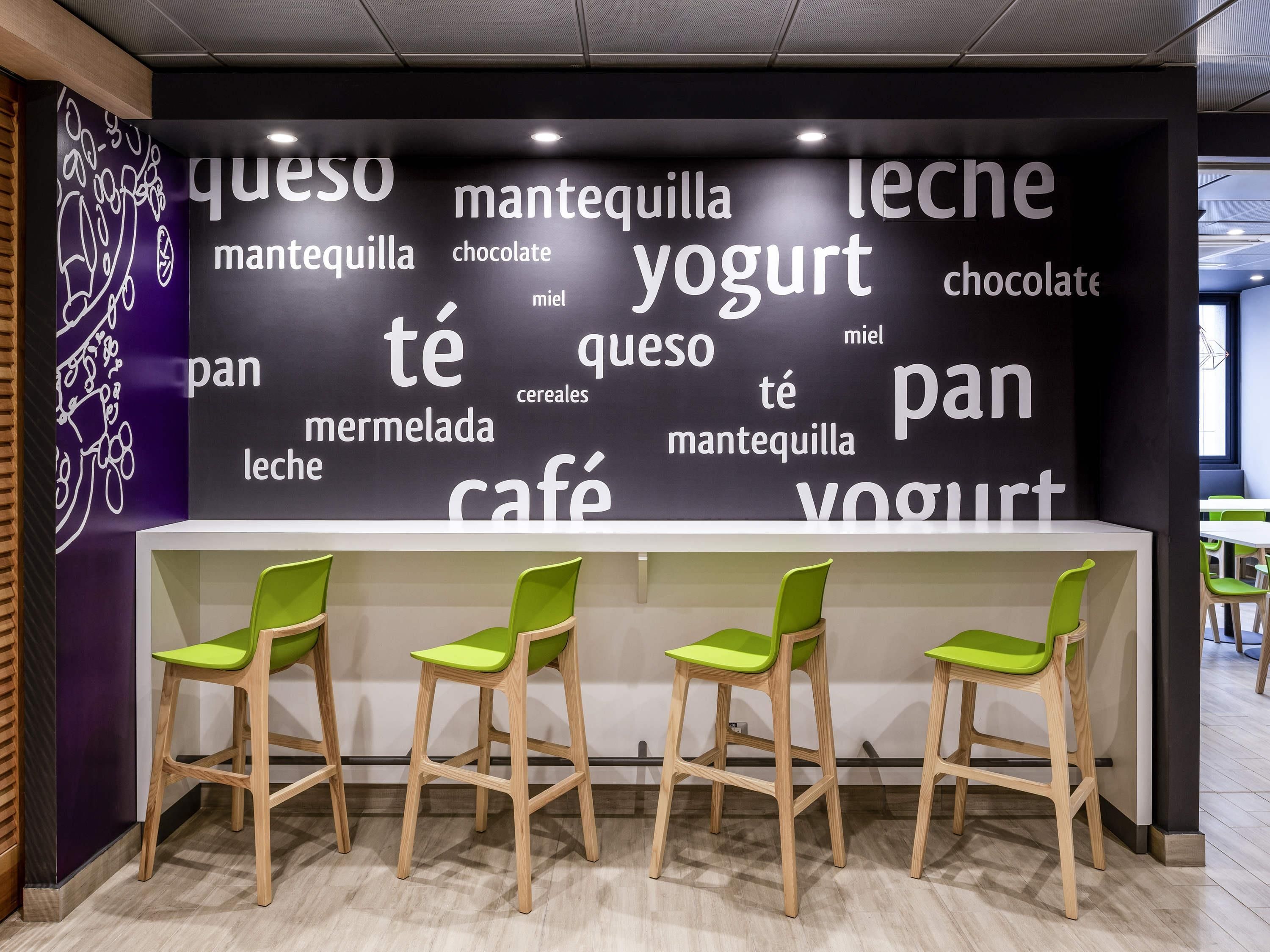 Una cafetería con una pared que muestra palabras como 'queso', 'mantequilla', 'leche', 'yogurt', 'pan', 'café' y 'cereales'. Hay tres sillas verdes frente a una barra blanca.