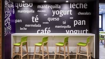 Un caffè con un muro che mostra parole come 'queso', 'mantequilla', 'leche', 'yogurt', 'pan', 'café' e 'cereali'. Tre sedie verdi davanti a un bancone bianco.