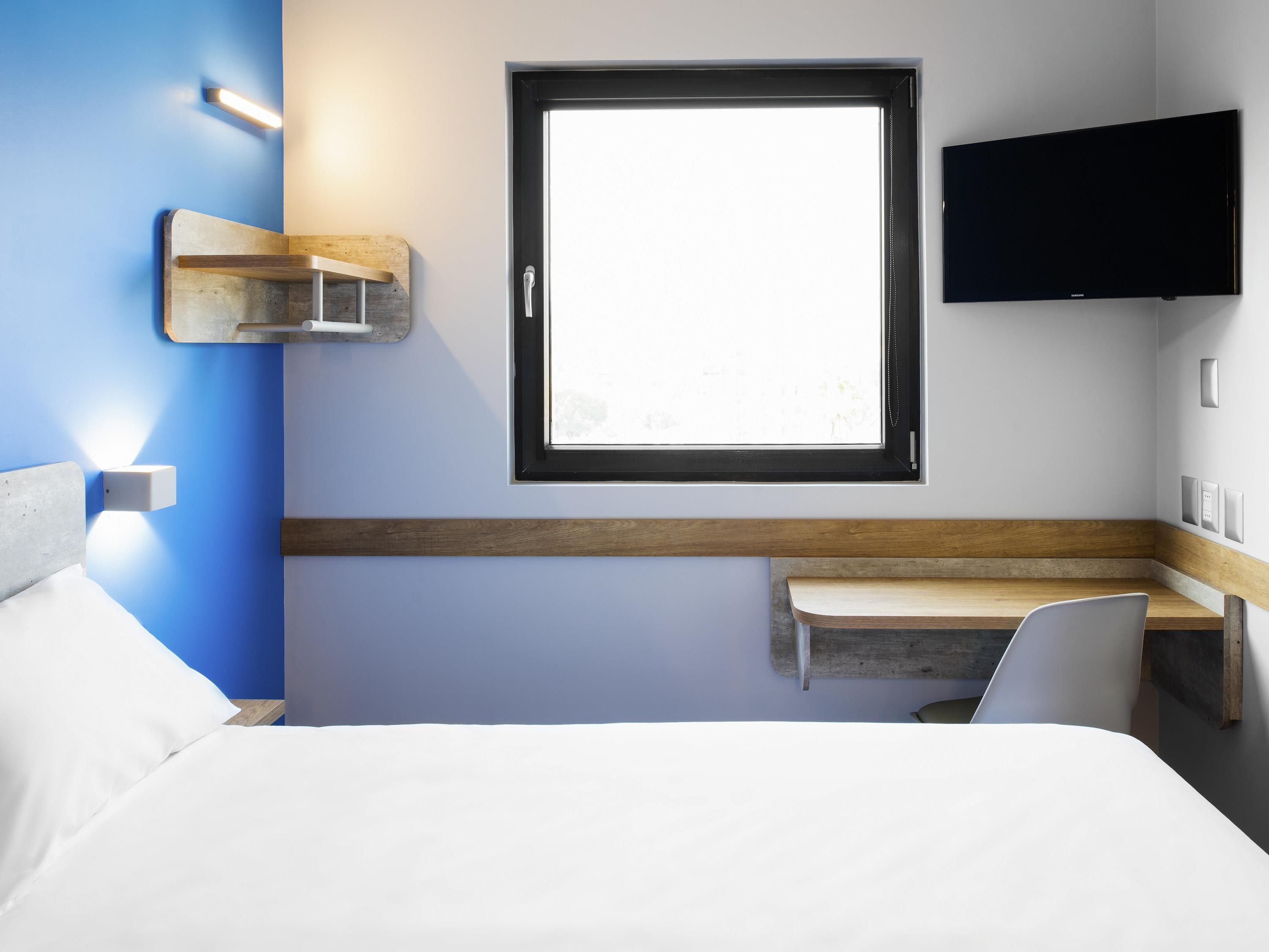 A hotel room with a bed, a desk, a window, a flat-screen TV, and a wall-mounted light fixture.