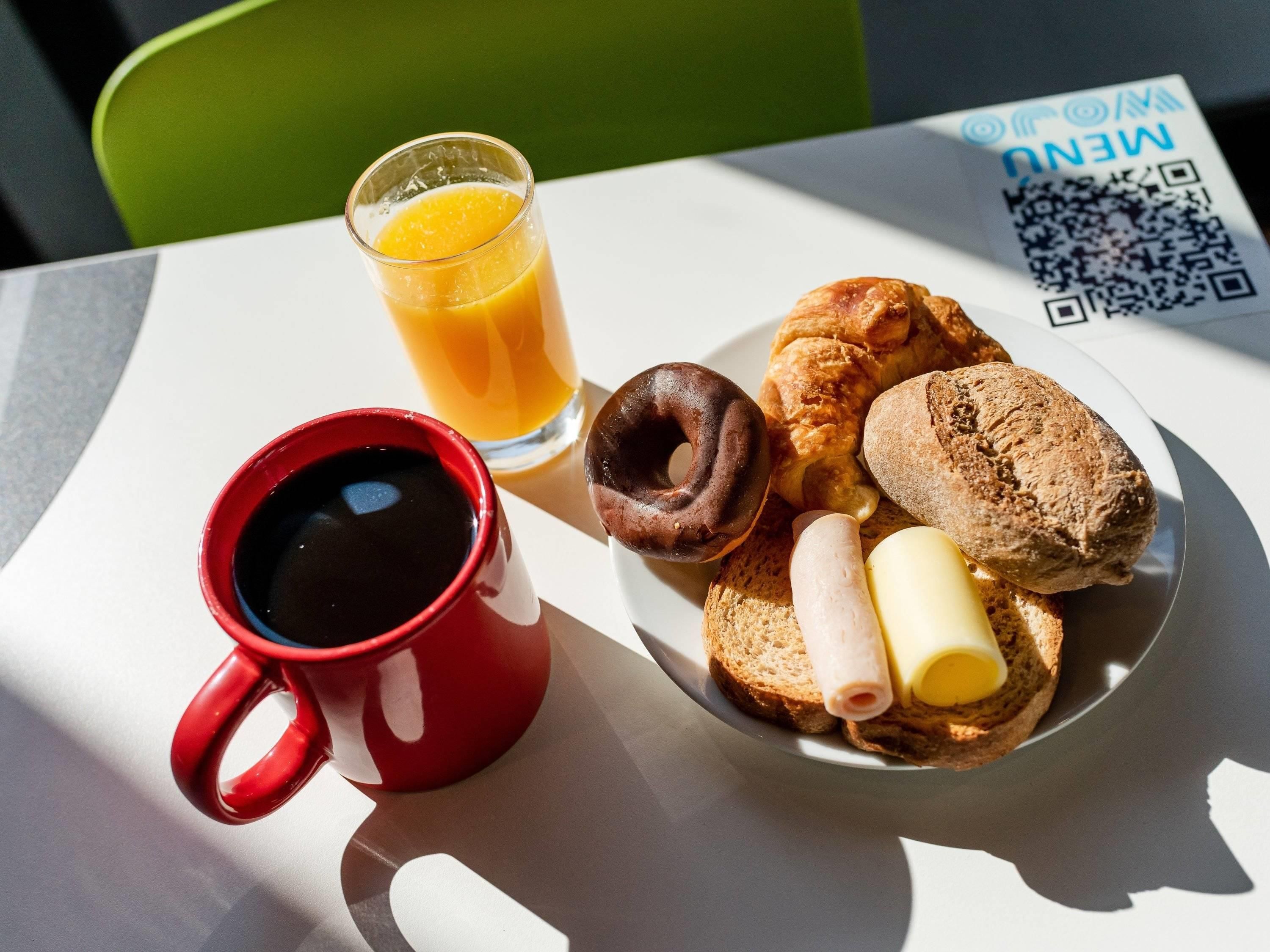 Un plato de desayuno con tostadas, un croissant, un donut de chocolate y un panecillo, acompañado de un vaso de jugo de naranja y una taza roja.
