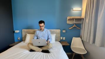 A person sitting on a bed with a laptop, in a room with a blue wall, a bedside table, and a hanging light fixture.