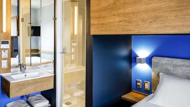 Moderni hotellihuone, jossa on lasiseinäinen suihkukaappi, puinen pesuallas ja sängyssä pääty.