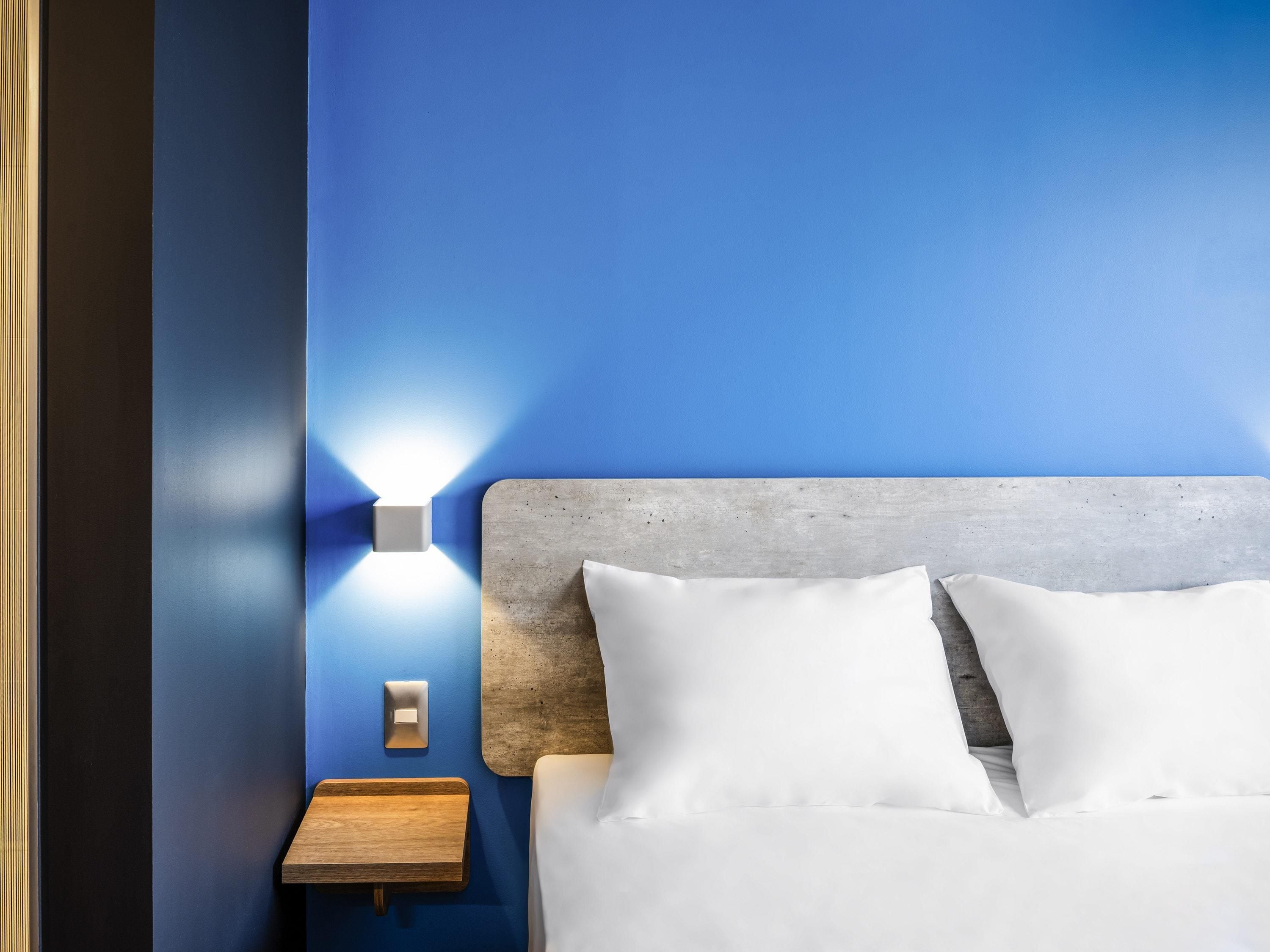 A hotel room with a blue wall, a wooden nightstand, a bed with white linens, and a wall-mounted light fixture.