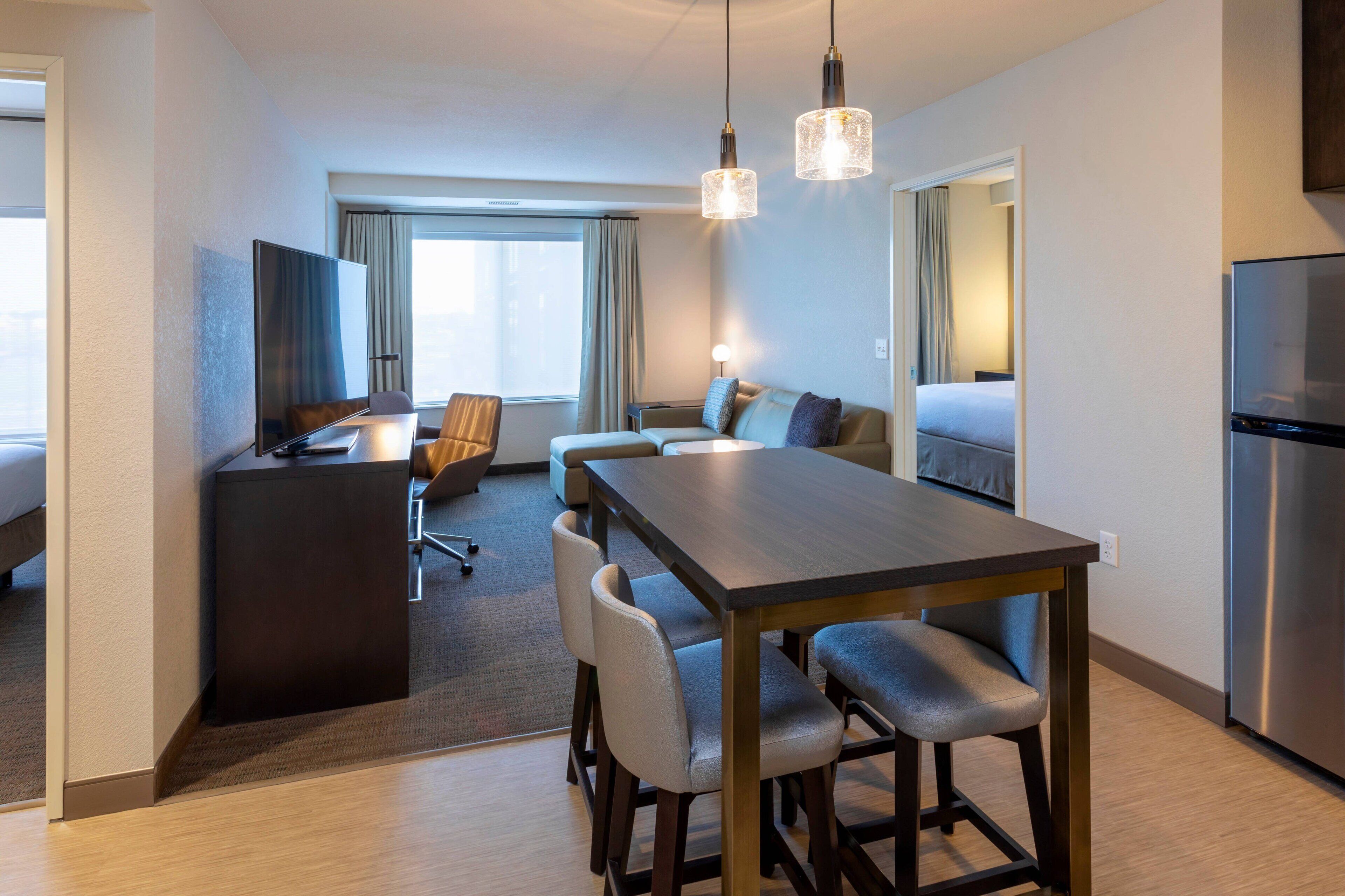 Una habitación de hotel moderna con zona de comedor, televisión, refrigerador y un amplio ventanal con cortinas.