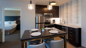 A modern hotel room with a dining area featuring a dark wooden table, two chairs, and a breakfast bar. There is a bed with a dark headboard, a small potted plant, and a nightstand with a lamp. The kitchen area includes a refrigerator, microwave, oven, and a coffee maker. - Residence Inn by Marriott Minneapolis Maple Grove/Arbor Lakes (Maple Grove)