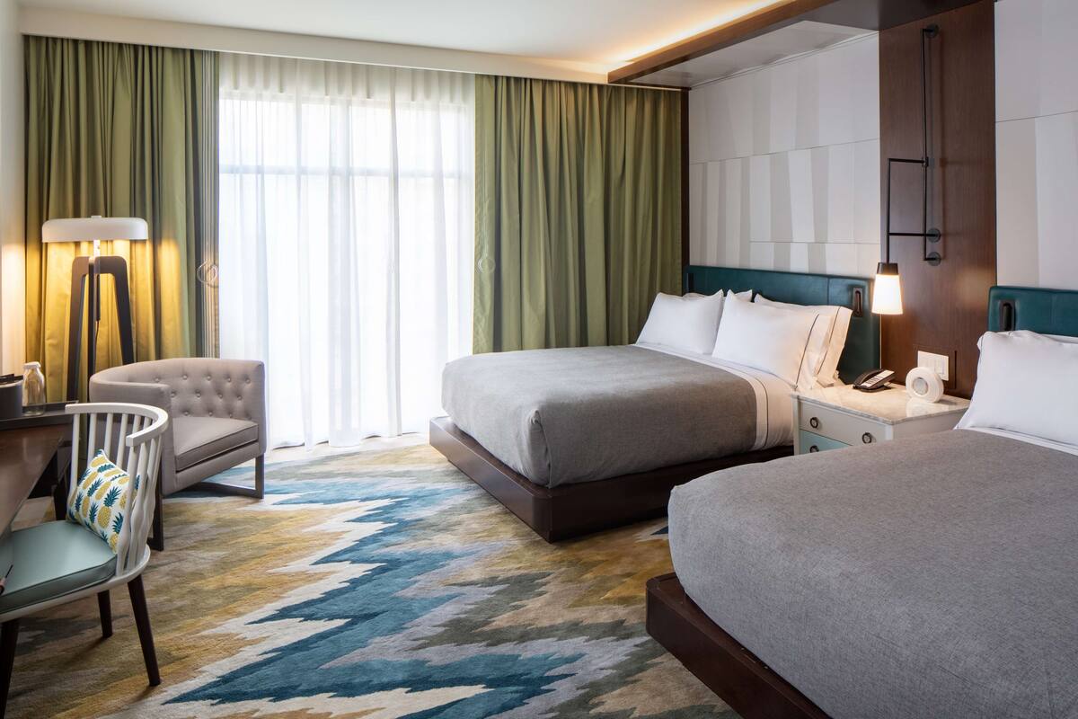Une chambre d’hôtel avec deux lits, un bureau avec une chaise, une lampe et une grande fenêtre avec des rideaux.