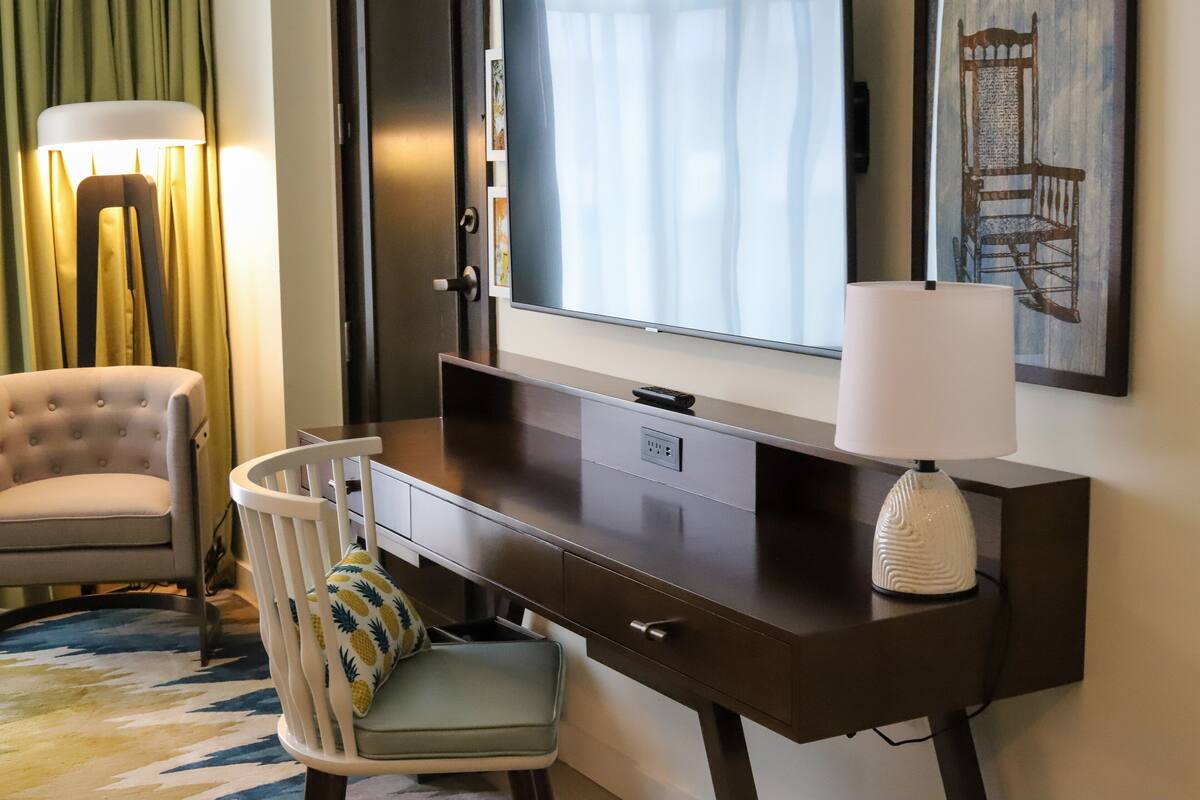 Une chambre d’hôtel dotée d’un grand téléviseur à écran plat fixé au mur, d’un bureau en bois avec une lampe, d’un fauteuil confortable et d’une petite table avec une chaise.