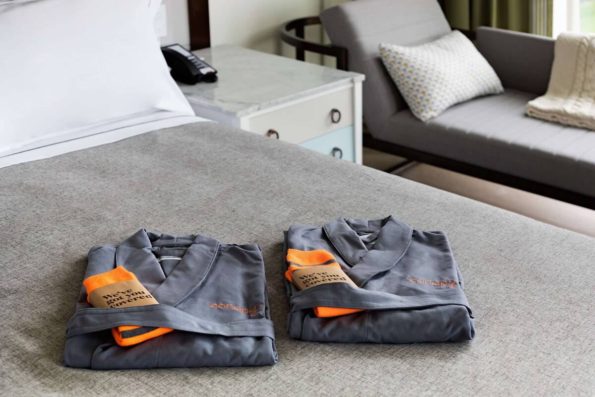 Deux peignoirs gris pliés, avec des étiquettes orange, sont posés sur un canapé gris dans une chambre d’hôtel.