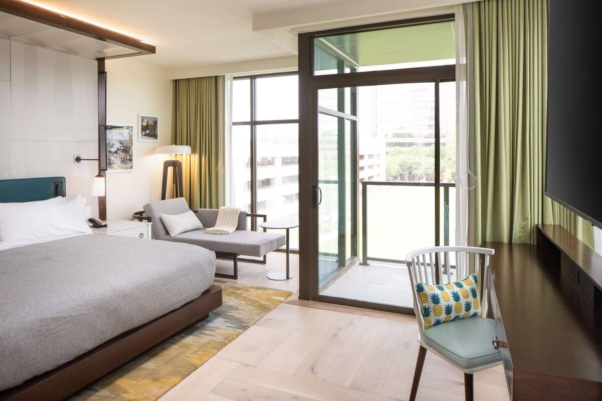 Une chambre d’hôtel avec un grand lit, un canapé, un fauteuil, une télévision et un balcon avec des rideaux.