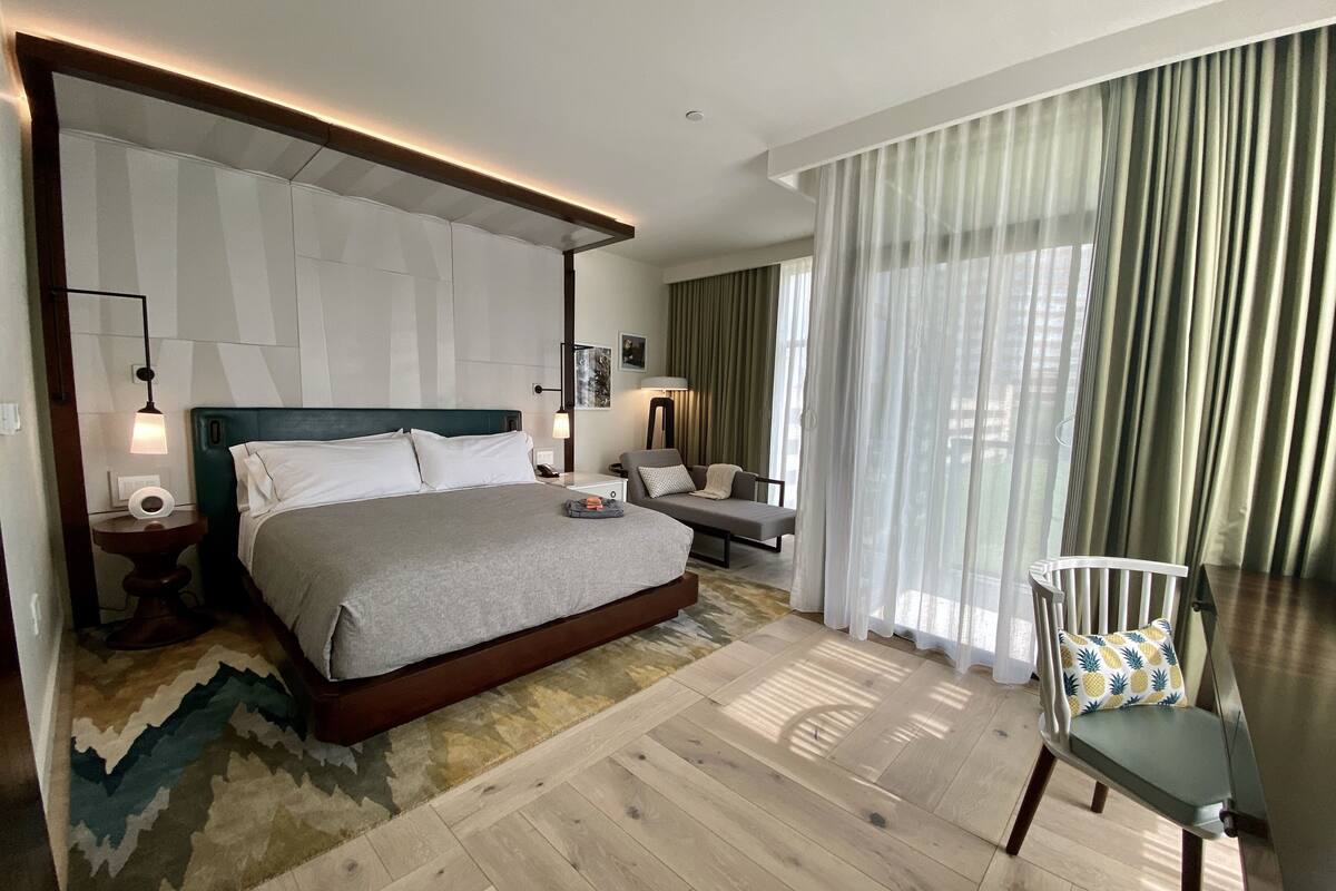 Une chambre d’hôtel moderne avec un grand lit, une table de chevet avec une lampe, un canapé, un fauteuil et une vue sur l’extérieur à travers des rideaux légers.