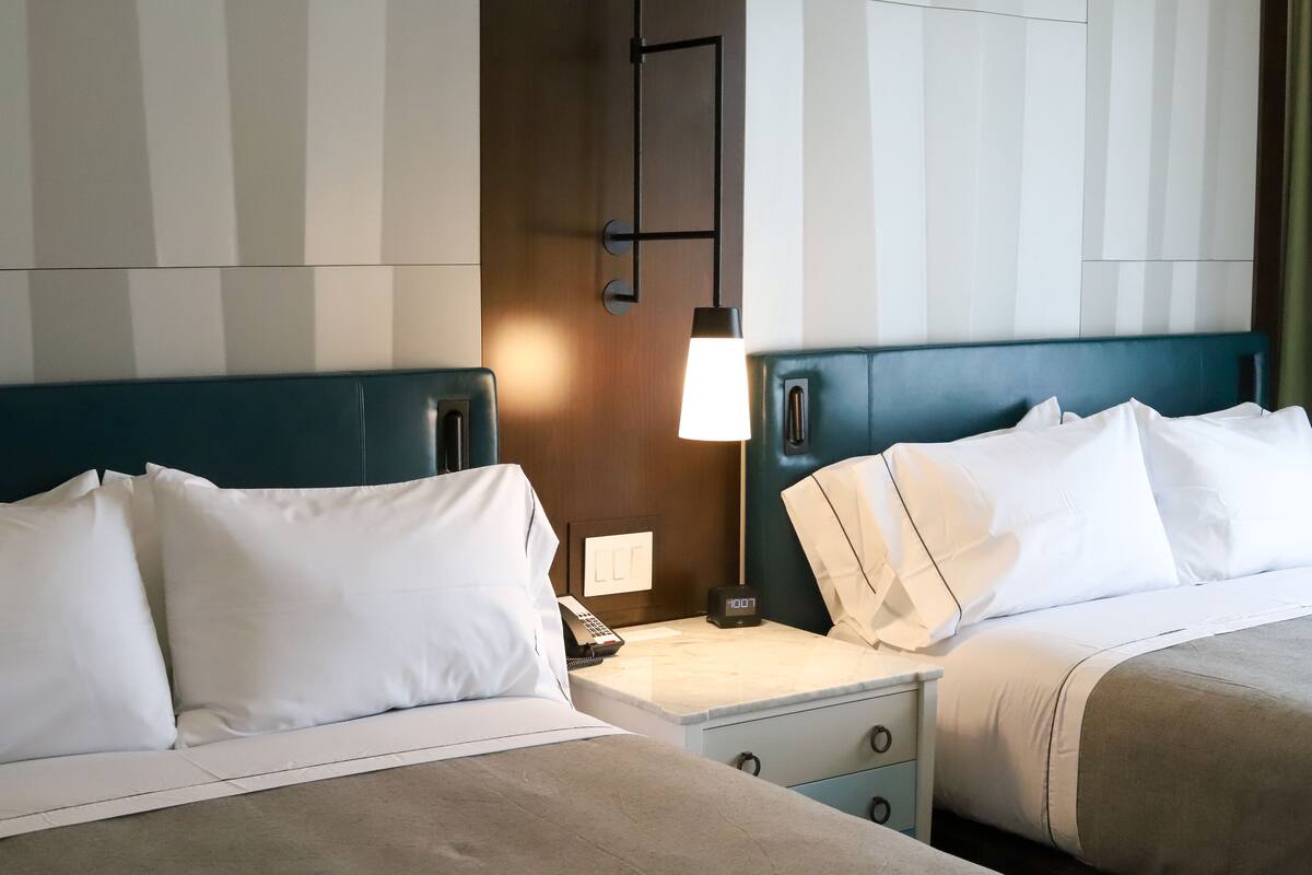 Une chambre d’hôtel avec deux lits, chacun doté de draps blancs et d’une tête de lit turquoise. On y trouve une table de chevet avec une lampe, un téléphone et une horloge numérique.