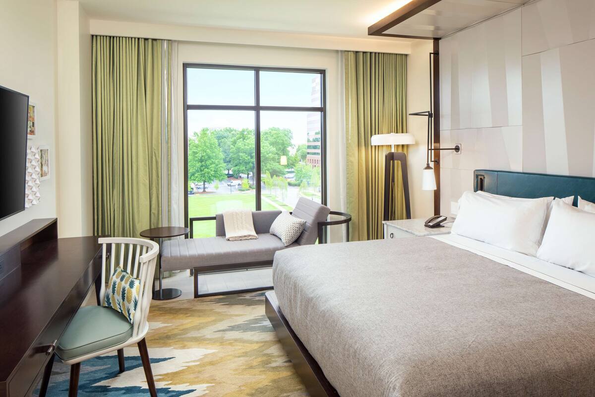 Une chambre d’hôtel avec un grand lit, un bureau, une chaise, une petite table et une fenêtre avec des rideaux.