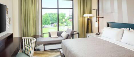 A hotel room with a large bed, a desk, a chair, a small table, and a window with curtains.