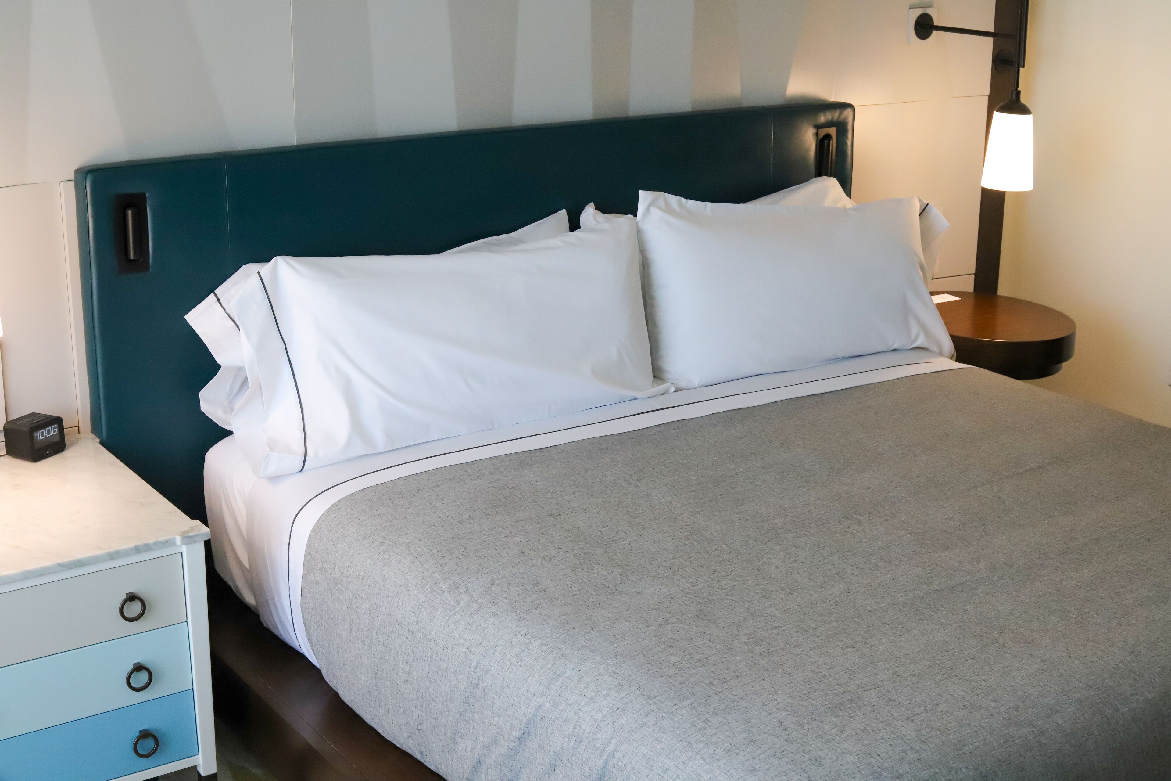 A neatly made bed with white bedding and a grey throw, a blue headboard, a bedside table with a lamp, and a small clock.