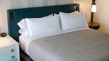 A neatly made bed with white bedding and a grey throw, a blue headboard, a bedside table with a lamp, and a small clock.