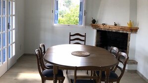 A dining area with a round wooden table, six chairs, a hanging pendant light, a window with shutters, and a fireplace with a mantel. - La Maison Bleu (Messini)