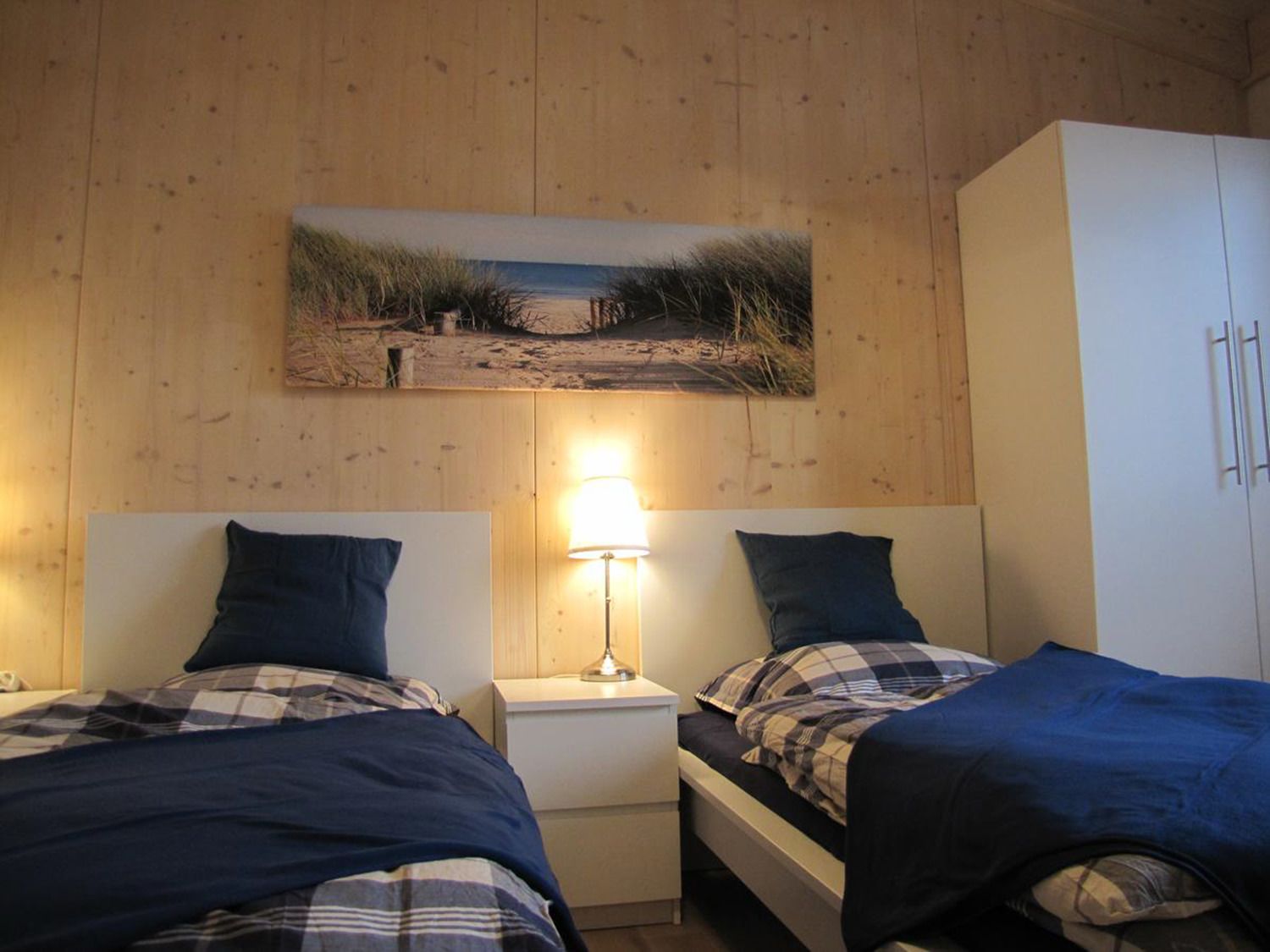 A room with two beds, a nightstand with a lamp, a wardrobe, and a beach-themed wall art.