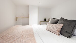 Un lit bien fait, recouvert d’une couverture blanche et rose, une table de chevet avec une plante, et un luminaire moderne.
