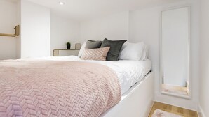 Une chambre à coucher moderne avec un lit blanc, une couverture rose à texture et une petite table de chevet agrémentée d’une plante.