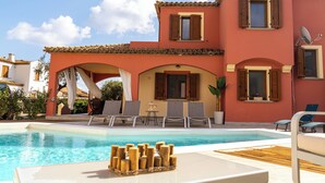 Un espace aménagé au bord de la piscine, avec des chaises longues, une table en bois et un bâtiment doté d’arcades et de fenêtres.