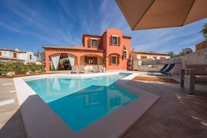 A poolside area with a clear blue pool, lounge chairs, and a building with a red facade.