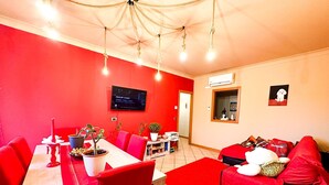 A dining area with a red table and chairs, a television mounted on the wall, and a sofa with red cushions.
