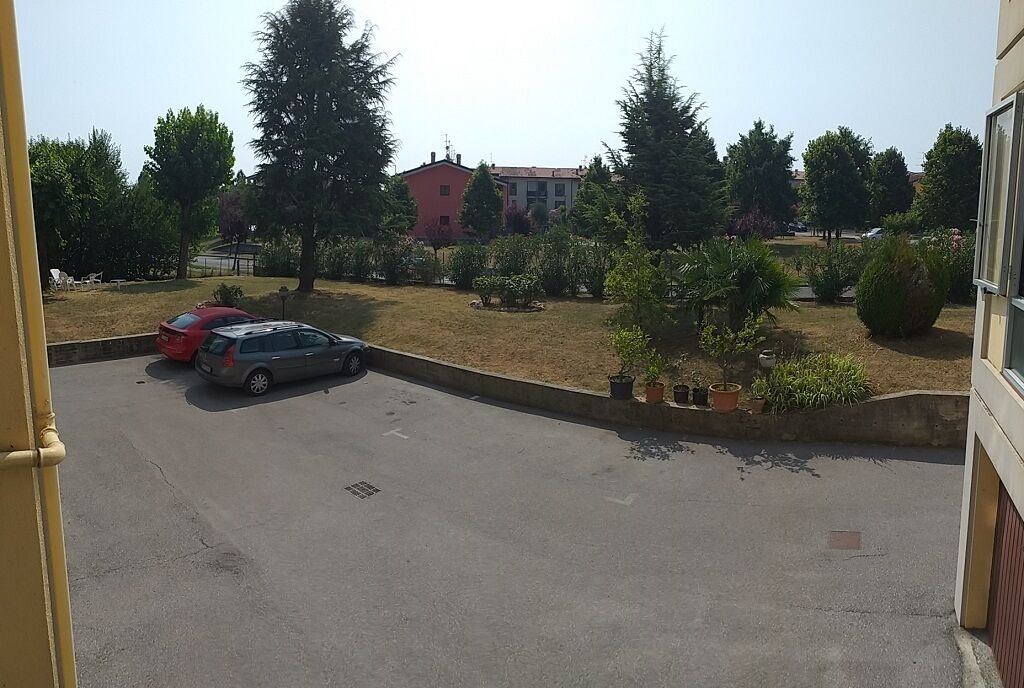Ein Parkplatz mit zwei Autos, ein Gebäude mit roter Fassade und eine Grünfläche mit Bäumen und Sträuchern.