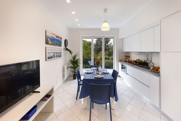 A modern kitchen with white cabinets, a dining area with a blue tablecloth, and a TV on a white stand.