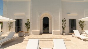Ein Gebäude mit einem zentralen Bogen, weißen Wänden und einer gepflasterten Terrasse mit Liegestühlen und Topfpflanzen.