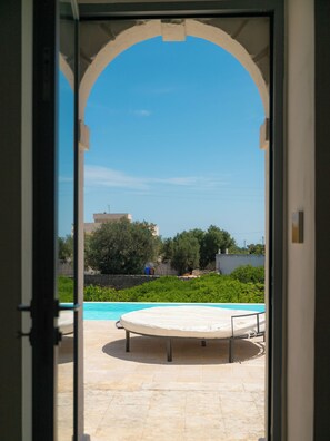 An outdoor swimming pool with a sun lounger, surrounded by greenery and a clear sky.