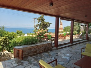 A covered outdoor seating area with stone flooring, wooden furniture, and a view of the sea.