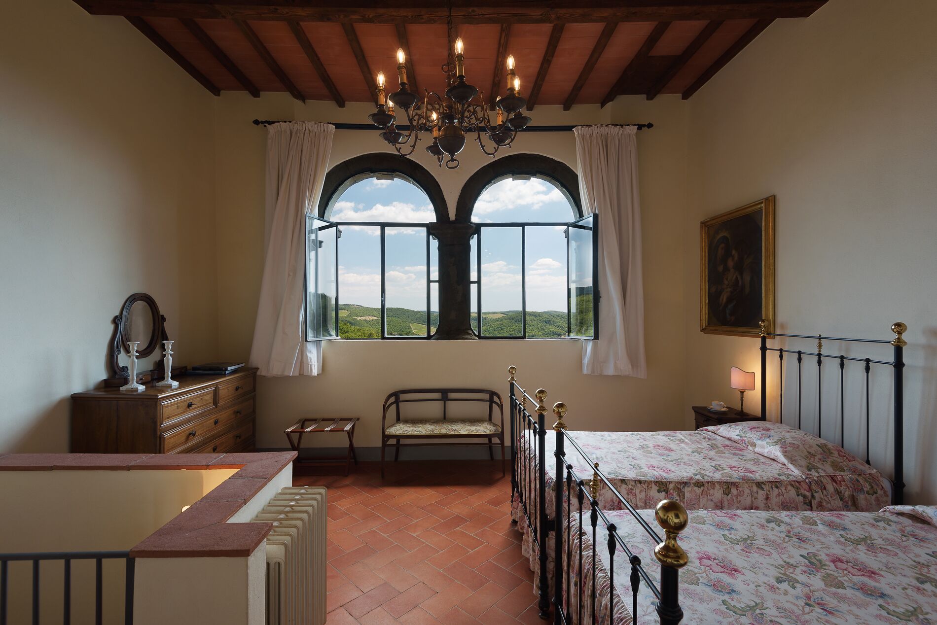 Une chambre avec une grande fenêtre en arc de cercle, un lit recouvert d’une couvre-lit à motifs floraux, une table de chevet avec une lampe et une commode en bois avec un miroir.