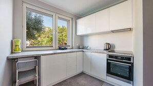 A modern kitchen with white cabinets, a built-in oven, and a window with a view of trees. - Apartamenty Sun & Snow Kopernika (Ustka)