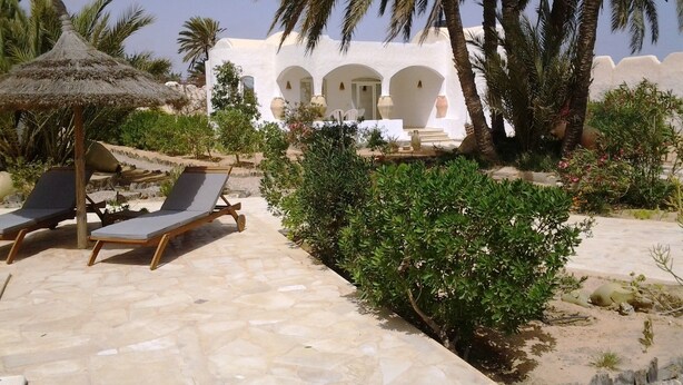 Resort com pátio de pedra, cadeiras de descanso e um edifício branco com arcos.
