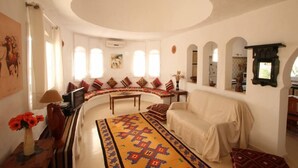 Quarto com cama sinuosa, sofá, televisão e mesa pequena. Há janelas com cortinas, ar condicionado fixado na parede e um espelho decorativo.