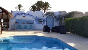 Piscina com parede revestida de azulejos representando um barco e arcos, mobiliário de exterior e palmeiras.