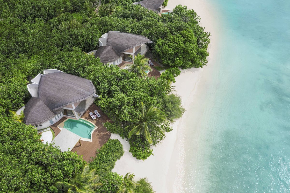 Vue aérienne d’un complexe hôtelier comprenant plusieurs bungalows sur pilotis, une piscine et une végétation luxuriante.