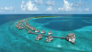 Une vue aérienne de bungalows sur pilotis reliés par des passerelles, entourés d’eaux turquoise et sous un ciel dégagé.