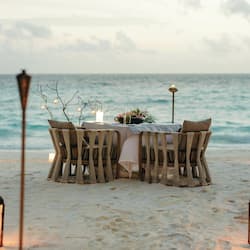 Enjoy a romantic beachfront dining experience with a table set for two, surrounded by wicker chairs and a beautiful floral arrangement.