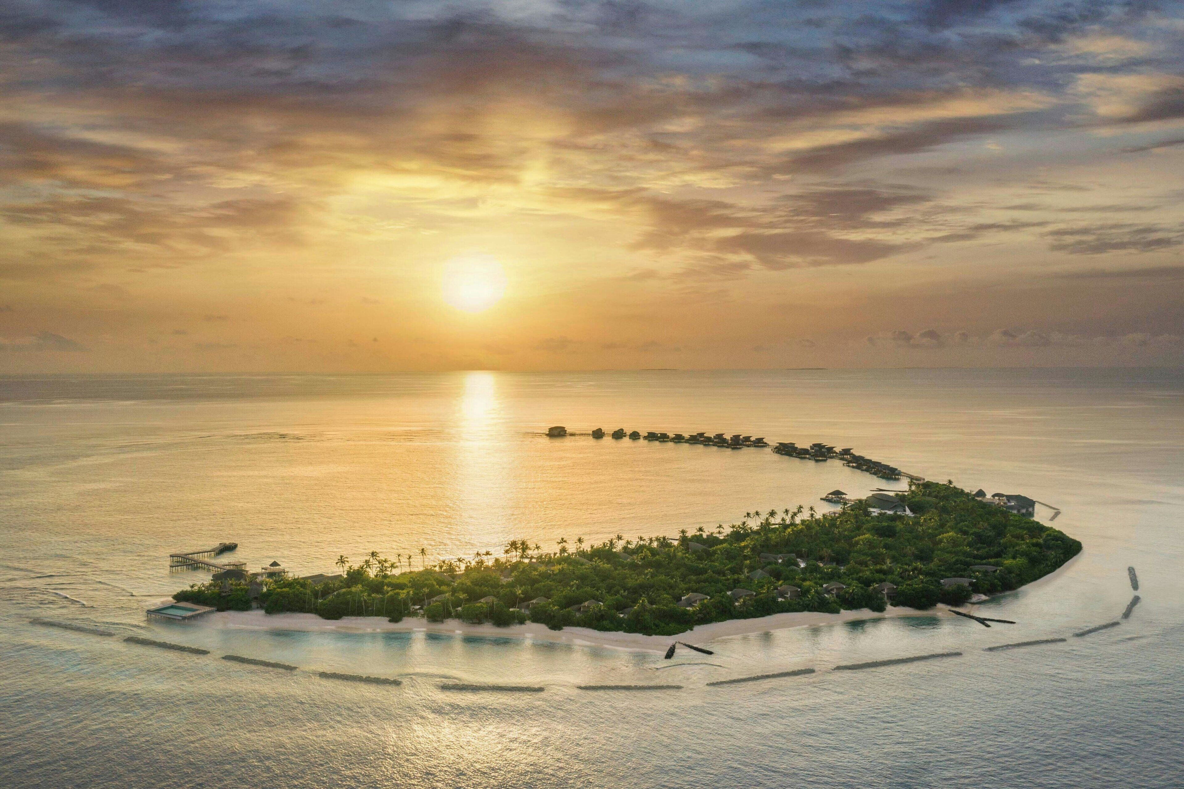 An aerial view of a resort island with a long pier, surrounded by water, and a sunset in the background.