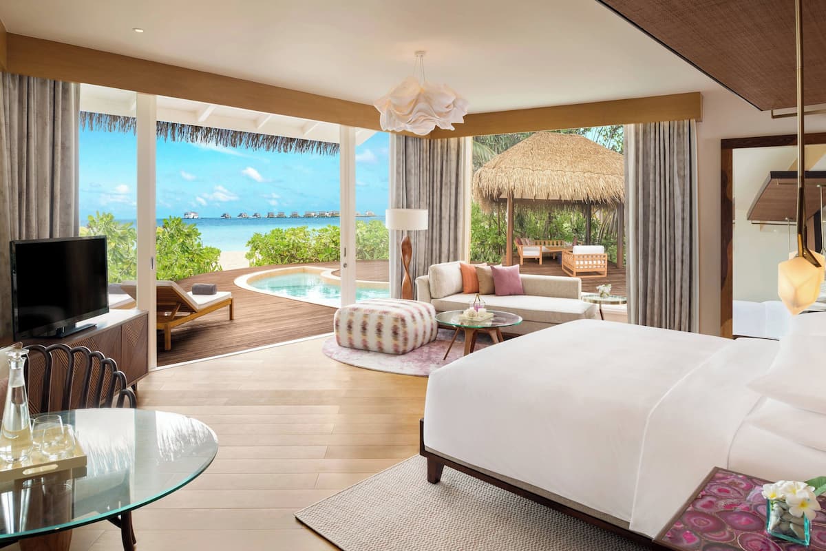 Une chambre spacieuse avec un grand lit, une table en verre et un téléviseur. Elle dispose d’un balcon donnant sur une piscine, d’une structure à toit de chaume et offre une vue sur la mer.