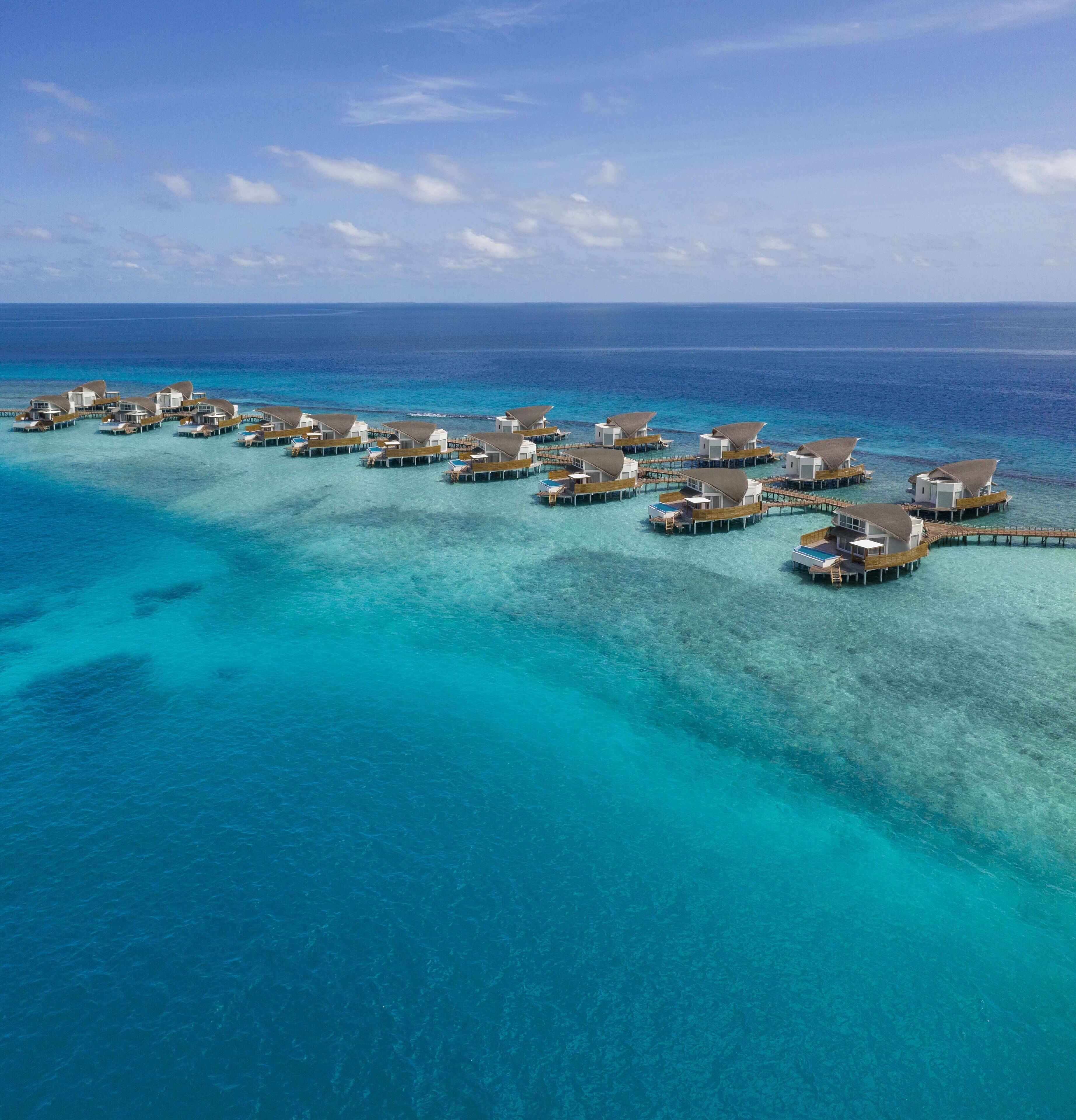 An aerial view of overwater bungalows connected by wooden walkways, set in clear turquoise waters with a calm sea and a clear sky.