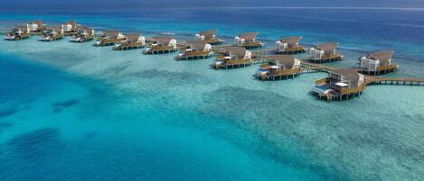 An aerial view of overwater bungalows connected by wooden walkways, set in clear turquoise waters with a calm sea and a clear sky.